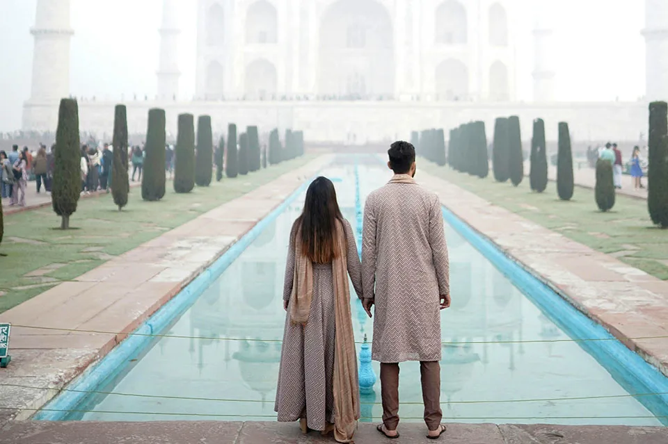 A couple dressed in traditional Indian attire holding hands and facing the Taj Mahal from the reflecting pool pathway on a foggy morning.