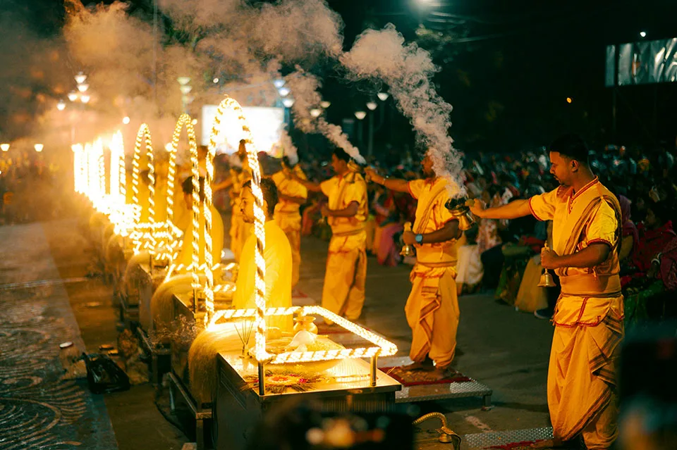 Men in traditional saffron attire perform the Ganga Aarti ritual at night, holding incense burners that release smoke, with a row of ornate lamps lit brightly in the foreground, and a large crowd seated behind them.