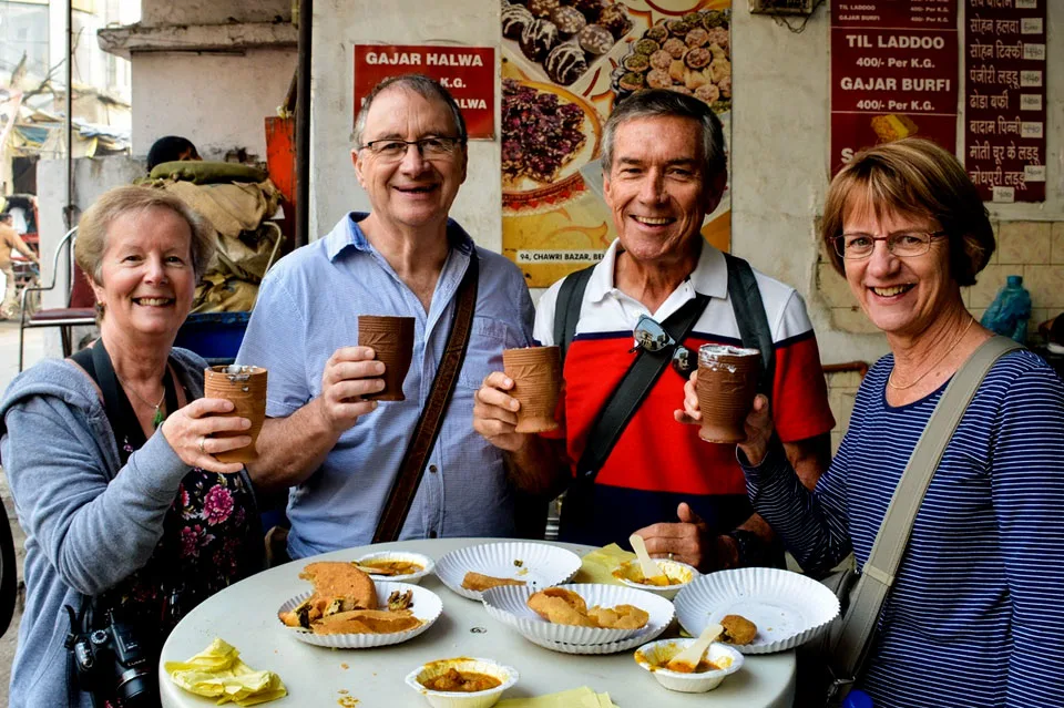 Four smiling tourists holding clay cups and enjoying Indian street food at a local shop in Delhi, with plates of snacks like samosas and sweets on the table.