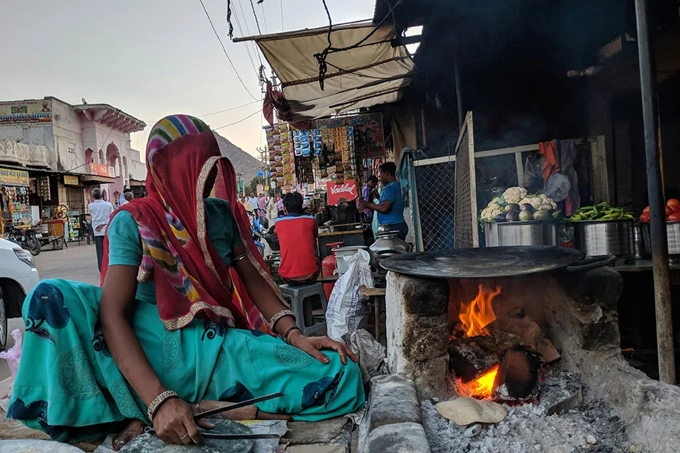 A woman in colorful traditional attire cooking on an open wood fire in a street market in Jaipur India, with vegetables and local vendors visible in the background.